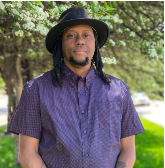Color Photo of Shawn Trenell O'Neal wearing a purple shirt and black hat standing  in front of a tree